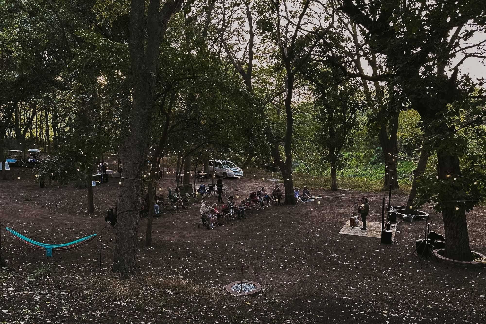An evening concert in the woods at Still Pastures: a small audience in camp chairs, a musician on a small stage, and string lights hung through the trees